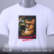 Футболка мотаюсь по бізнесу, білого кольору, розкладена на поверхні, фон світлий, принт з коргі в окулярах, що сидить за кермом, з текстом "Мотаюсь по бізнесу!" червоного кольору, на футболці є етикетка з зеленим шнурком.