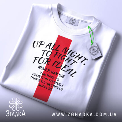 Футболка Мотиваційна Цитата. Біла футболка, складена на поверхні, з червоним прямокутником на грудях, на якому написані мотиваційні фрази чорним шрифтом. На футболці є етикетка з зеленим шнурком. Фон світлий, однорідний.