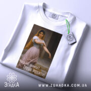 Футболка мотивуюча дівчина, біла, складена на поверхні, фон світлий, принт з дівчиною в традиційному вбранні, текст на українській мові: 'Чого тут думати? Треба робити!', на ярлику зелена стрічка.