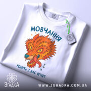 Футболка мовчання робить ягнят. Біла футболка з принтом, що зображає яскравого лева, розташована на плоскій поверхні. Принт яскравий, з оранжевими та зеленими відтінками, текст на українській мові. Фон світло-сірий, футболка має етикетку з зеленим шнурком.