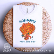 Футболка мовчання робить ягнят. Біла футболка з принтом, що зображає яскравого лева, розташована на плетеній кошику. Принт яскравий, з оранжевими та зеленими відтінками, текст на українській мові. Фон світло-сірий, футболка акуратно складена.