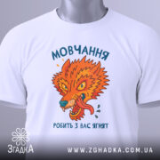 Футболка мовчання робить ягнят. Біла футболка з принтом, що зображає яскравого лева, розташована на плоскій поверхні. Принт яскравий, з оранжевими та зеленими відтінками, текст на українській мові. Фон світло-сірий, футболка має етикетку з зеленим шнурком.