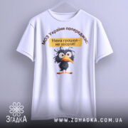 Футболка МОЗ України попереджає: біла футболка, підвішена на вішалці, з принтом чорного птаха та текстом, на фоні світлого кольору.