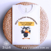 Футболка МОЗ України попереджає: складена біла футболка з принтом чорного птаха, текстом зверху, на фоні плетеного кошика.