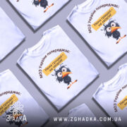 Футболка МОЗ України попереджає: кілька складених білих футболок з принтом чорного птаха, текстом зверху, на фоні світло-сірого кольору.