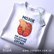 Футболка мозок другий орган. Біла футболка, складена вдвічі, з принтом червоного мозку та рукою, що вказує на нього. Фон світлий, зображення з верхнього ракурсу. На футболці є етикетка з зеленим шнурком.