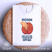 Футболка мозок другий орган. Біла футболка, складена на плетеній основі, з принтом червоного мозку та рукою, що вказує на нього. Фон нейтральний, зображення з верхнього ракурсу. Текст на футболці чорного кольору.