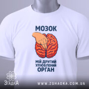 Футболка мозок другий орган. Біла футболка, складена вдвічі, з принтом червоного мозку та рукою, що вказує на нього. Фон світлий, зображення з верхнього ракурсу. На футболці є етикетка з зеленим шнурком.