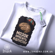 Футболка мозок просить відпустку. Біла футболка, складена на поверхні, з принтом мозку під парасолькою. Ракурс зверху, фон світлий. На футболці видно етикетку з зеленим шнурком.