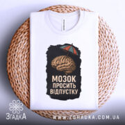 Футболка мозок просить відпустку. Біла футболка, складена на плетеній основі, з принтом мозку під парасолькою. Ракурс зверху, фон світлий. Принт яскравий, з чіткими деталями.