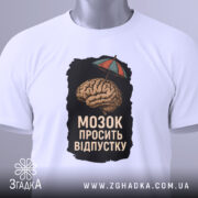 Футболка мозок просить відпустку. Біла футболка, складена на поверхні, з принтом мозку під парасолькою. Ракурс зверху, фон світлий. На футболці видно етикетку з зеленим шнурком.