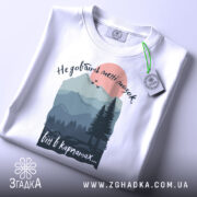 Футболка мозок в Карпатах, біла, складена на столі, фон світлий, принт з горами, лісом і сонцем, текст зверху: 'Не добивай метті мозок', внизу: 'віт в карпатах...', з етикеткою на рукаві.