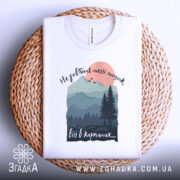 Футболка мозок в Карпатах, біла, складена на плетеній основі, фон світлий, принт з горами, лісом і сонцем, текст зверху: 'Не добивай метті мозок', внизу: 'віт в карпатах...'.