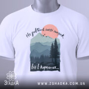 Футболка мозок в Карпатах, біла, складена на столі, фон світлий, принт з горами, лісом і сонцем, текст зверху: 'Не добивай метті мозок', внизу: 'віт в карпатах...', з етикеткою на рукаві.