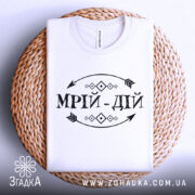 Футболка мрій дій, білого кольору, складена на плетеній основі, фон світло-сірий, принт чорного кольору з написом 'МРІЙ-ДІЙ' та декоративними елементами.