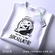 Футболка мрій і досліджуй, біла, складена на столі, фон світлий, принт з космічним кораблем та місяцем, текст 'Досліджуй!' під малюнком, з зеленим ярликом.