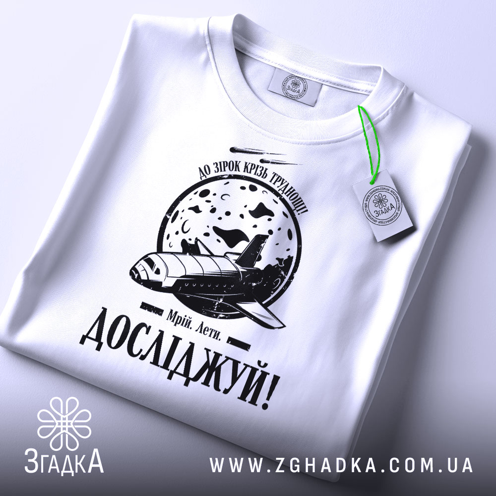 Футболка мрій і досліджуй, біла, складена на столі, фон світлий, принт з космічним кораблем та місяцем, текст 'Досліджуй!' під малюнком, з зеленим ярликом.