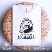 Футболка мрій і досліджуй, біла, складена на плетеній основі, фон світлий, принт з космічним кораблем та місяцем, текст 'Досліджуй!' під малюнком.