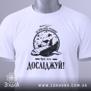 Футболка мрій і досліджуй, біла, складена на столі, фон світлий, принт з космічним кораблем та місяцем, текст 'Досліджуй!' під малюнком, з зеленим ярликом.