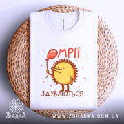 Футболка мрії здуваються, біла, зображена зверху на плетеній кошику, фон світло-сірий, принт з жовтим сонечком, що тримає червоний повітряний шар, текст 'ОМПІІ ЗДУВАЮТЬСЯ' у червоному кольорі.