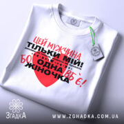 Футболка мужчина мій, біла, складена на столі, фон світлий, принт червоний та чорний з текстом, на футболці є етикетка.