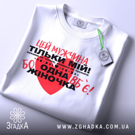Футболка мужчина мій, біла, складена на столі, фон світлий, принт червоний та чорний з текстом, на футболці є етикетка.
