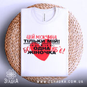 Футболка мужчина мій, біла, складена на плетеній кошику, фон нейтральний, принт червоний та чорний з текстом, що містить серце.