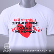Футболка мужчина мій, біла, складена на столі, фон світлий, принт червоний та чорний з текстом, на футболці є етикетка.