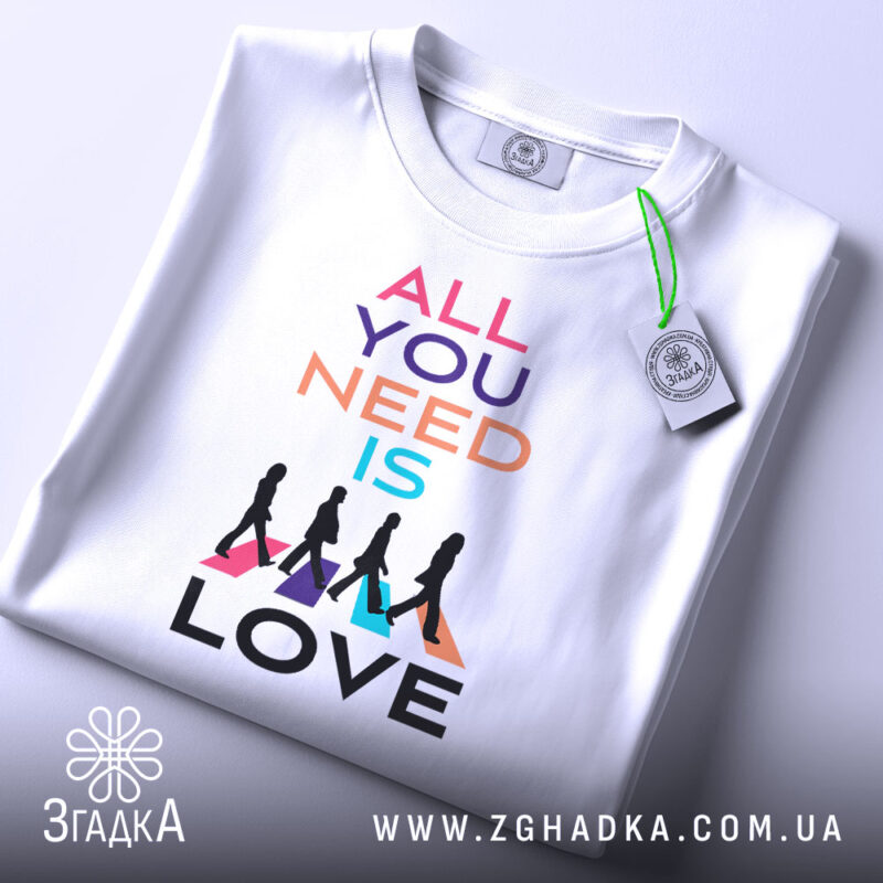 Футболка музика серця. Біла футболка, складена на столі, фон світло-сірий. Принт з текстом 'ALL YOU NEED IS LOVE' у яскравих кольорах, з прикріпленою етикеткою зеленого кольору.