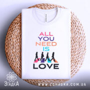 Футболка музика серця. Біла футболка, складена на плетеній кошику, фон світло-сірий. Принт містить текст 'ALL YOU NEED IS LOVE' у різних кольорах, а також силуети людей, що йдуть, на кольорових трикутниках.