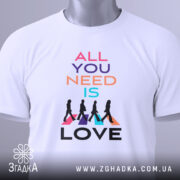 Футболка музика серця. Біла футболка, складена на столі, фон світло-сірий. Принт з текстом 'ALL YOU NEED IS LOVE' у яскравих кольорах, з прикріпленою етикеткою зеленого кольору.