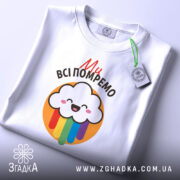 Футболка ми всі помремо, біла, зображена в складеному вигляді, фон світло-сірий, принт з хмаринкою та кольоровими краплями, на ярлику зелена стрічка.