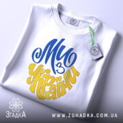 Футболка ми з України білого кольору, зображена зверху, складена на поверхні, фон світло-сірий, принт у синьому та жовтому кольорах, напис "Ми з України" розташований по центру, на футболці є етикетка з зеленим шнурком.