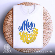 Футболка ми з України білого кольору, зображена зверху, складена на плетеній кошику, фон світло-сірий, принт у синьому та жовтому кольорах, напис "Ми з України" розташований по центру.