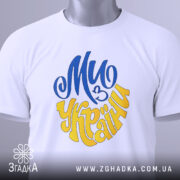 Футболка ми з України білого кольору, зображена зверху, складена на поверхні, фон світло-сірий, принт у синьому та жовтому кольорах, напис "Ми з України" розташований по центру, на футболці є етикетка з зеленим шнурком.