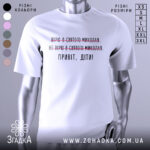 Футболка Миколай дітям. Білий колір, зображена з фронтального ракурсу на манекені. Фон світло-сірий, принт містить текст з червоними закресленнями та чорним написом 'Привіт, діти!'.