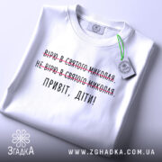 Футболка Миколай дітям. Білий колір, зображена в складеному вигляді на світлому фоні. Принт містить текст з червоними закресленнями та чорним написом 'Привіт, діти!', з зеленим етикеткою.