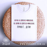 Футболка Миколай дітям. Білий колір, зображена в складеному вигляді на плетеній основі. Принт містить текст з червоними закресленнями та чорним написом 'Привіт, діти!'.