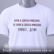 Футболка Миколай дітям. Білий колір, зображена в складеному вигляді на світлому фоні. Принт містить текст з червоними закресленнями та чорним написом 'Привіт, діти!', з зеленим етикеткою.
