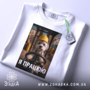 Футболка мила робота, білого кольору, зображена в складеному вигляді, фон світлий, принт з собачкою в жовтій касці, текст "Я ПРАЦЮЮ" під зображенням, на футболці є етикетка з зеленим шнурком.