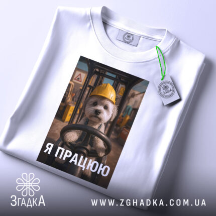 Футболка мила робота, білого кольору, зображена в складеному вигляді, фон світлий, принт з собачкою в жовтій касці, текст "Я ПРАЦЮЮ" під зображенням, на футболці є етикетка з зеленим шнурком.