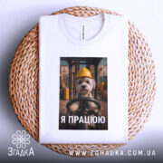 Футболка мила робота, білого кольору, складена на плетеній кошику, фон світлий, принт з собачкою в жовтій касці, текст "Я ПРАЦЮЮ" під зображенням, видно етикетку на горловині.