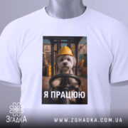 Футболка мила робота, білого кольору, зображена в складеному вигляді, фон світлий, принт з собачкою в жовтій касці, текст "Я ПРАЦЮЮ" під зображенням, на футболці є етикетка з зеленим шнурком.