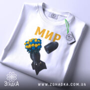 Футболка мир без війни, біла, складена на поверхні, фон світлий, принт з квітами жовтого та синього кольору на чорній бомбі, текст 'МИР' золотистого кольору, прикріплена етикетка.