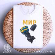 Футболка мир без війни, біла, складена на кошику, фон нейтральний, принт з квітами жовтого та синього кольору на чорній бомбі, текст 'МИР' золотистого кольору.
