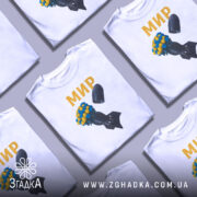Футболка мир без війни, біла, зображена на манекені, фон нейтральний, принт з квітами жовтого та синього кольору на чорній бомбі, текст 'МИР' золотистого кольору.