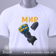 Футболка мир без війни, біла, складена на поверхні, фон світлий, принт з квітами жовтого та синього кольору на чорній бомбі, текст 'МИР' золотистого кольору, прикріплена етикетка.