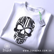 Футболка мисливець дух, біла, складена на поверхні, фон світло-сірий, принт чорного кольору з черепом та написом 'HUNTING MY HOBBY', з етикеткою зеленого кольору.
