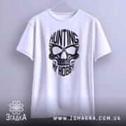 Футболка мисливець дух, біла, підвішена на вішалці, фон світло-сірий, принт чорного кольору з черепом та написом 'HUNTING MY HOBBY'.