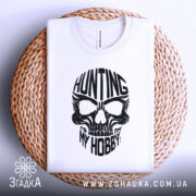 Футболка мисливець дух, біла, складена на плетеній кошику, фон світло-сірий, принт чорного кольору з черепом та написом 'HUNTING MY HOBBY'.